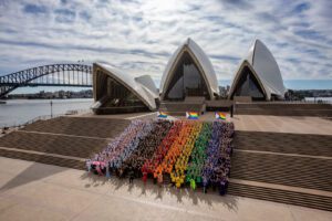 People are standing in front of Syndey, Australia's famous arches. They are wearing white, pink, blue, black red, orange, yellow, green, blue, and black again. They are standing in color block order to make up the Human Progress Flag.